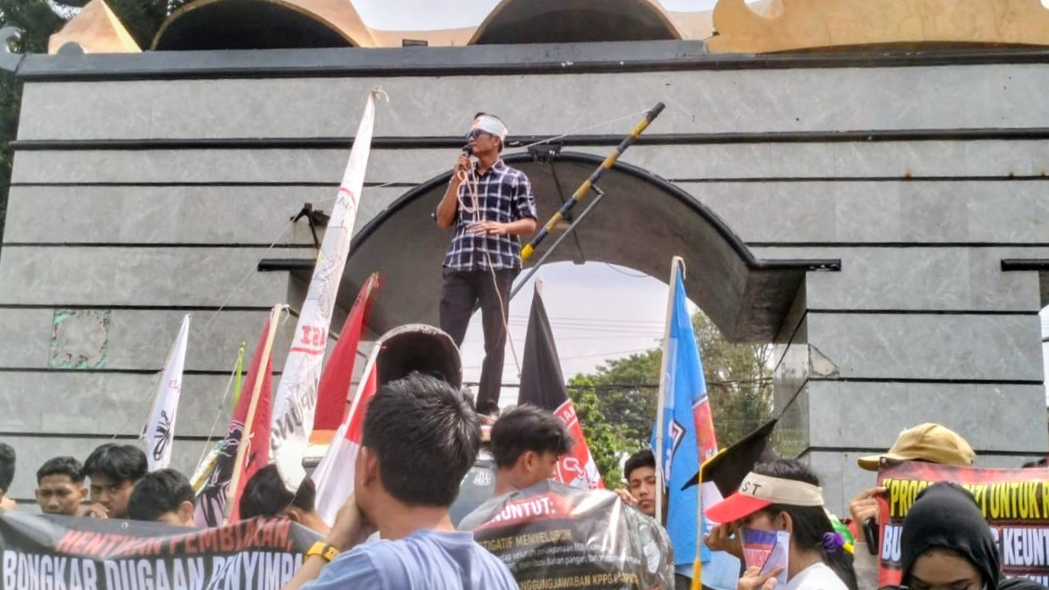 Seorang orator Aliansi Lampung Bersatu Awasi Kebijakan dan Anggaran berorasi menggunakan pengeras suara di depan gerbang Kantor DPRD Provinsi Lampung, Kamis 25/6/2026.