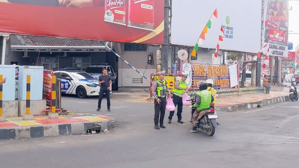 BANDAR LAMPUNG - Personel Pos Lantas Tugu Adipura Polresta Bandar Lampung membagikan paket makanan gratis kepada pengendara saat kegiatan Jumat Berkah di depan Pos Lantas Tugu Adipura, Jumat (17/7/2026). FOTO: PROYEKSI.ID